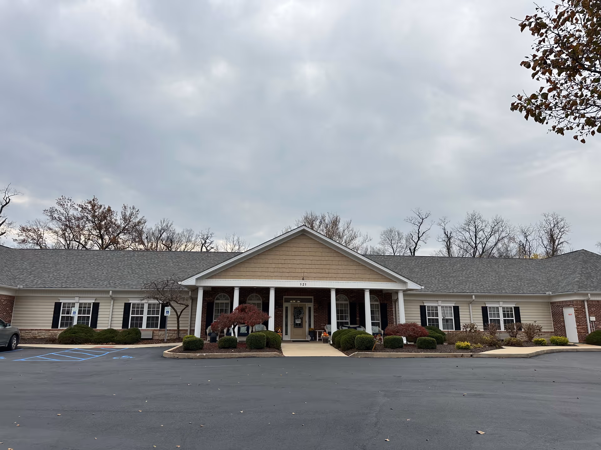 Front exterior view of a single-story senior living facility building with a covered entrance supported by white columns, landscaped bushes, and a paved parking area with handicap parking spaces. The sky is overcast and trees without leaves are visible in the background.