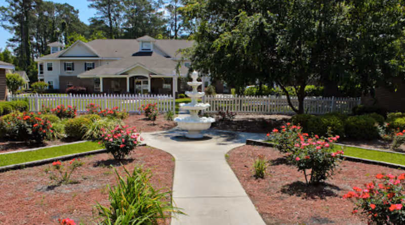 A landscaped outdoor garden area with a concrete pathway leading to a white multi-tiered fountain. The garden is bordered by flower beds with red flowers and green shrubs, surrounded by a white picket fence. In the background, there is a two-story building with a covered porch and trees providing shade.