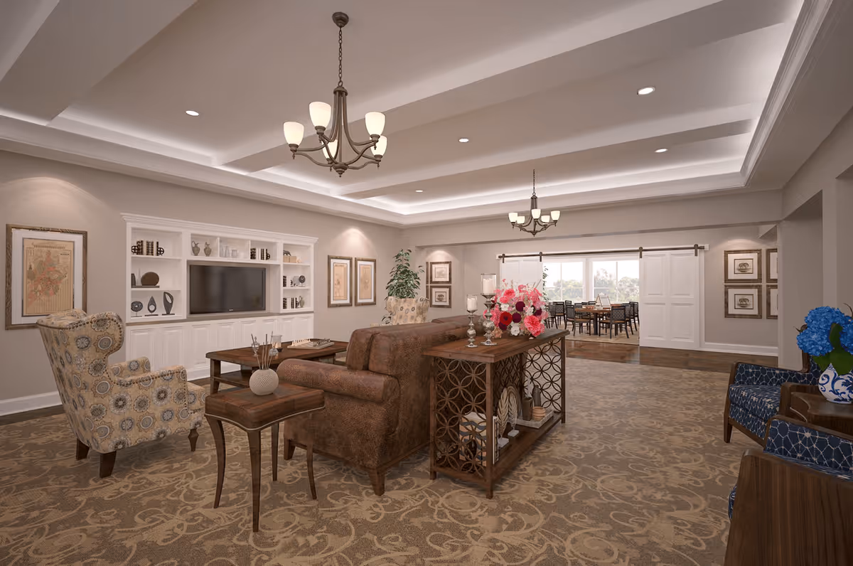 A spacious and elegantly decorated living room in a senior living facility featuring a brown leather sofa, patterned armchairs, a wooden coffee table, and a built-in white entertainment center with a TV and decorative items. The room has beige walls, framed artwork, a patterned carpet, and two chandeliers hanging from a tray ceiling. In the background, there is a dining area visible through a wide doorway with sliding barn doors.