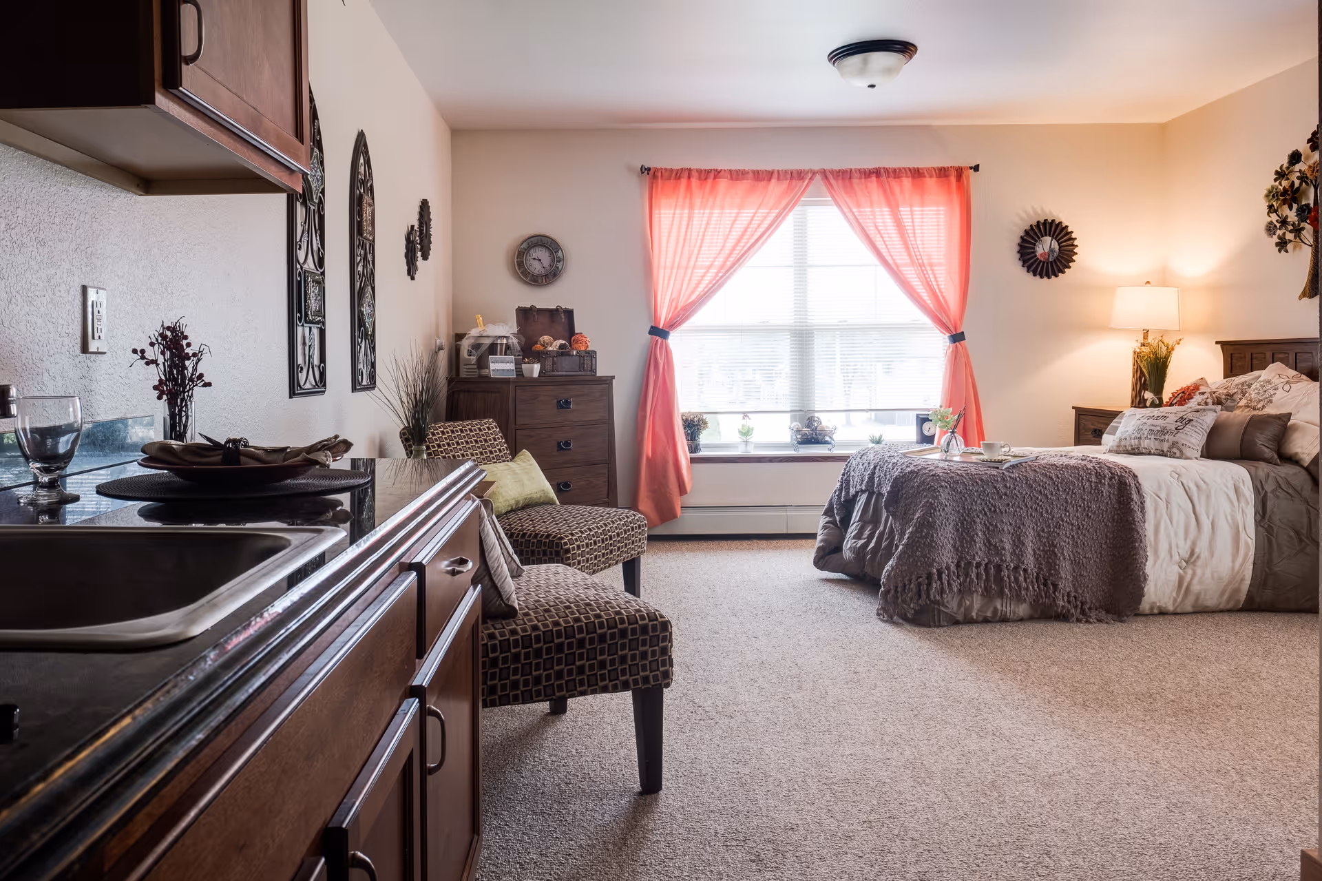 A cozy senior living facility room with a bed covered in a gray blanket and multiple pillows, a wooden nightstand with a lamp, two patterned chairs with a green pillow, a wooden dresser, and a large window with pink curtains letting in natural light. The foreground shows a kitchenette area with a sink and countertop.