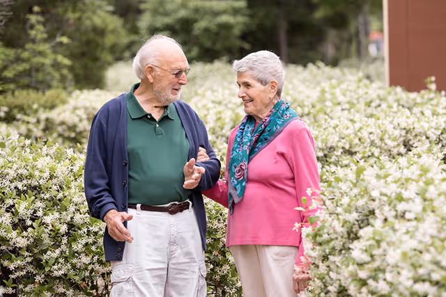 An elderly man and woman walking outdoors along a path lined with white flowering bushes, smiling and holding arms while engaging in conversation.