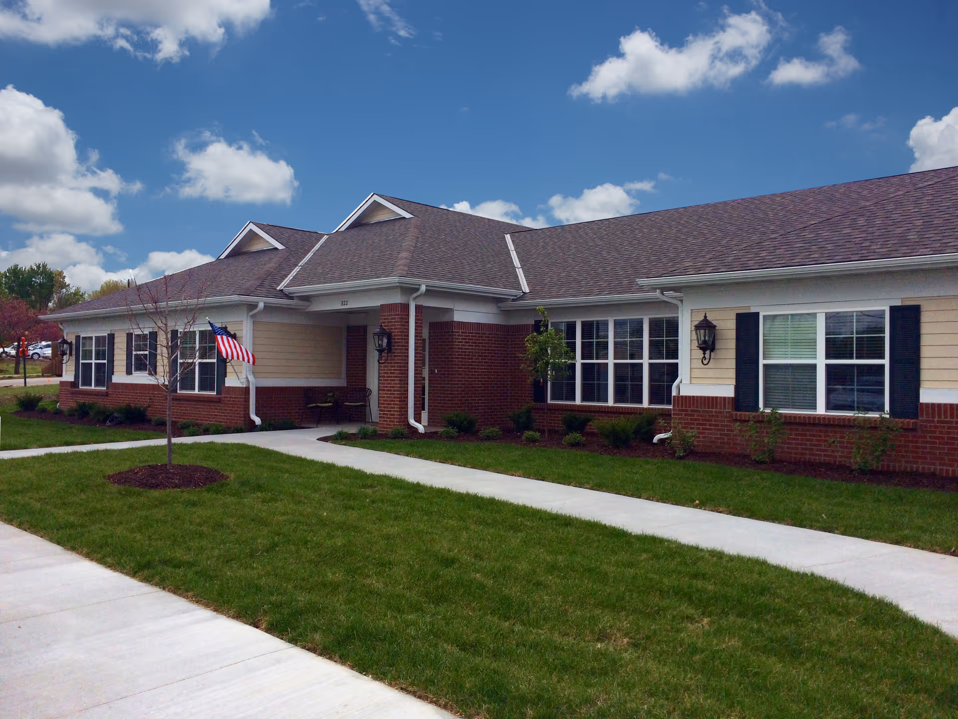 Exterior view of a single-story brick and siding building with a gabled roof under a partly cloudy sky. The building has multiple windows with black shutters, a small porch area with a bench, and an American flag mounted near the entrance. There is a well-maintained lawn with a young tree and concrete walkways leading to the entrance.
