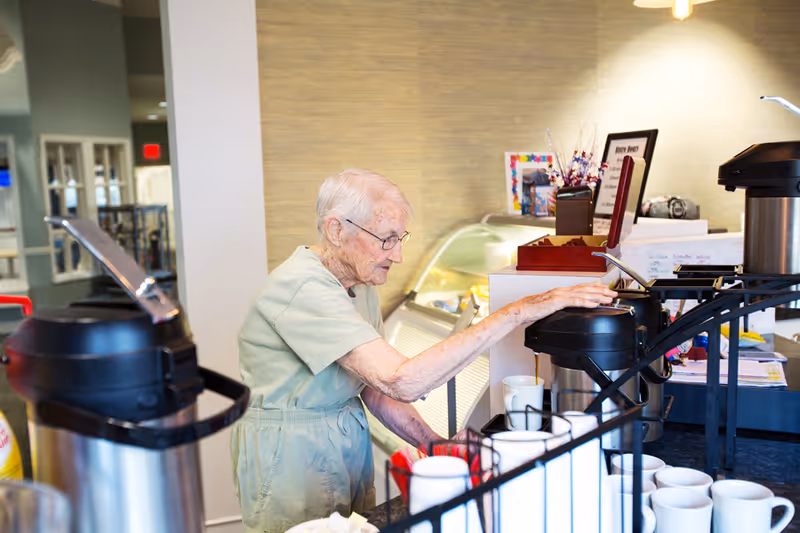 An elderly woman wearing glasses and a light green dress is reaching for a coffee dispenser in a well-lit room with a counter holding multiple coffee cups and condiments.