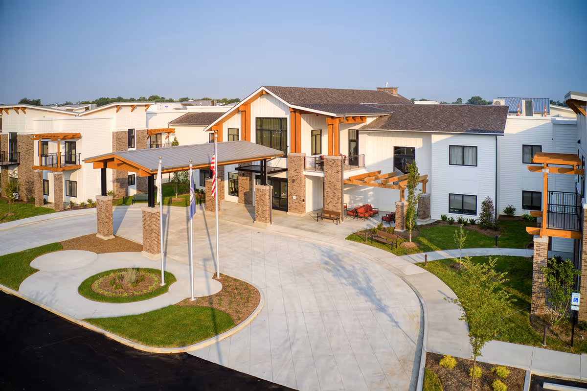 Exterior view of Preston Greens Senior Living facility showing a modern building with white siding, stone accents, and wooden beams. The entrance features a covered driveway with three flagpoles and landscaped greenery around the paved circular driveway.