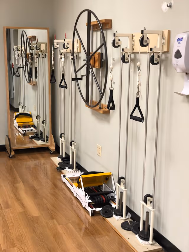 A rehabilitation exercise room with pulley systems mounted on the wall, a large wheel for arm exercises, weight plates on the floor, and a full-length mirror reflecting the equipment. A hand sanitizer dispenser is mounted on the wall.
