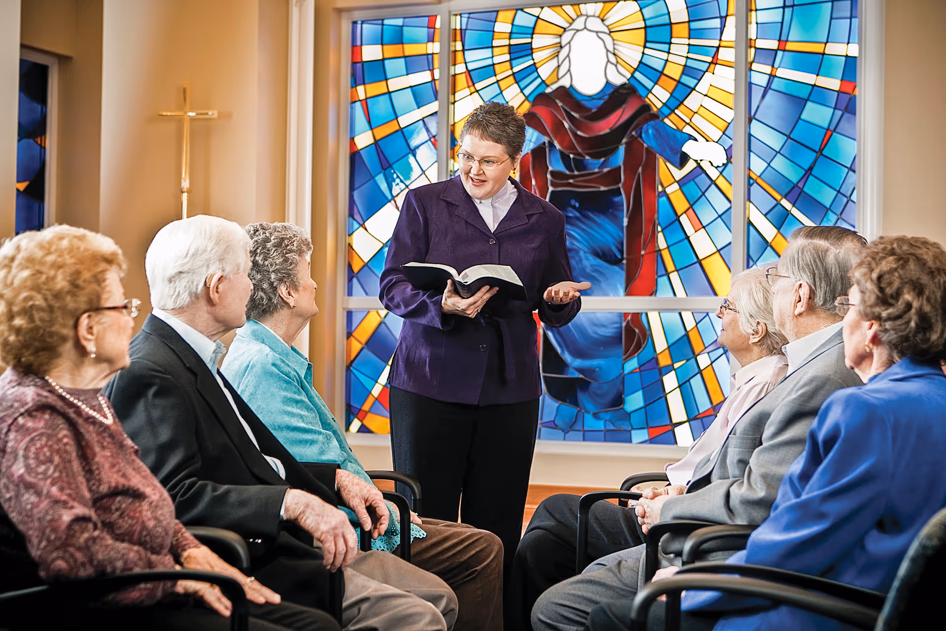 A woman holding an open book, possibly a Bible, is speaking to a group of elderly people seated in chairs arranged in a semi-circle. Behind her is a large colorful stained glass window depicting a religious figure with outstretched arms and a glowing halo. A gold cross is visible on the wall to the left.