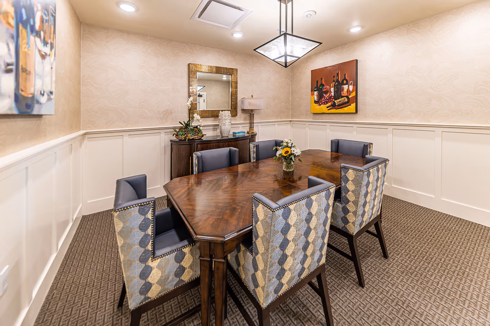 A well-lit dining room with a wooden dining table surrounded by six upholstered chairs with patterned backs. The room features beige textured wallpaper with white wainscoting, a decorative mirror above a sideboard, a table lamp, and two paintings on the walls. A small flower arrangement is placed in the center of the table.