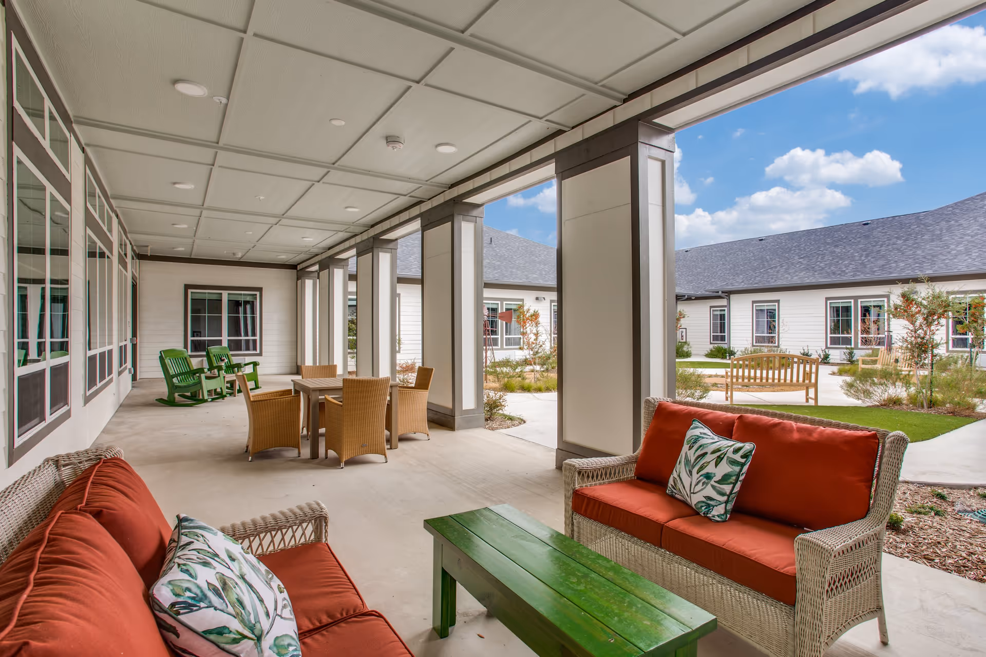 Covered outdoor patio area at Harvest of Aledo Senior Living with wicker furniture including two sofas with red cushions and leaf-patterned pillows, a green wooden coffee table, a dining table with four wicker chairs, and two green rocking chairs. The patio overlooks a courtyard with benches, landscaping, and a building with multiple windows under a partly cloudy sky.