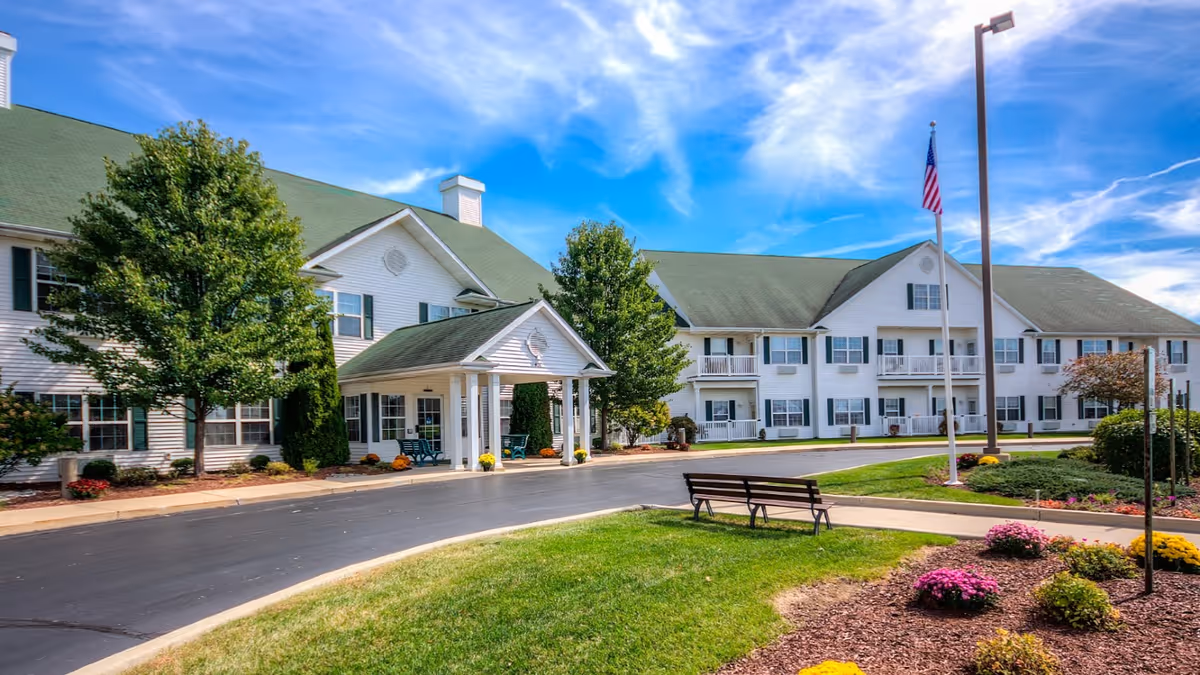 Exterior view of McKay Manor, a large two-story white building with green roof and multiple windows. The entrance has a covered porch with white pillars. There are trees, flower beds, a bench, and a flagpole with an American flag in the landscaped front area under a partly cloudy blue sky.