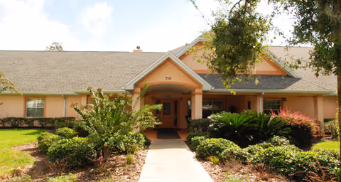 Front entrance of a single-story assisted living building with a covered portico, walkway, and landscaped shrubs.