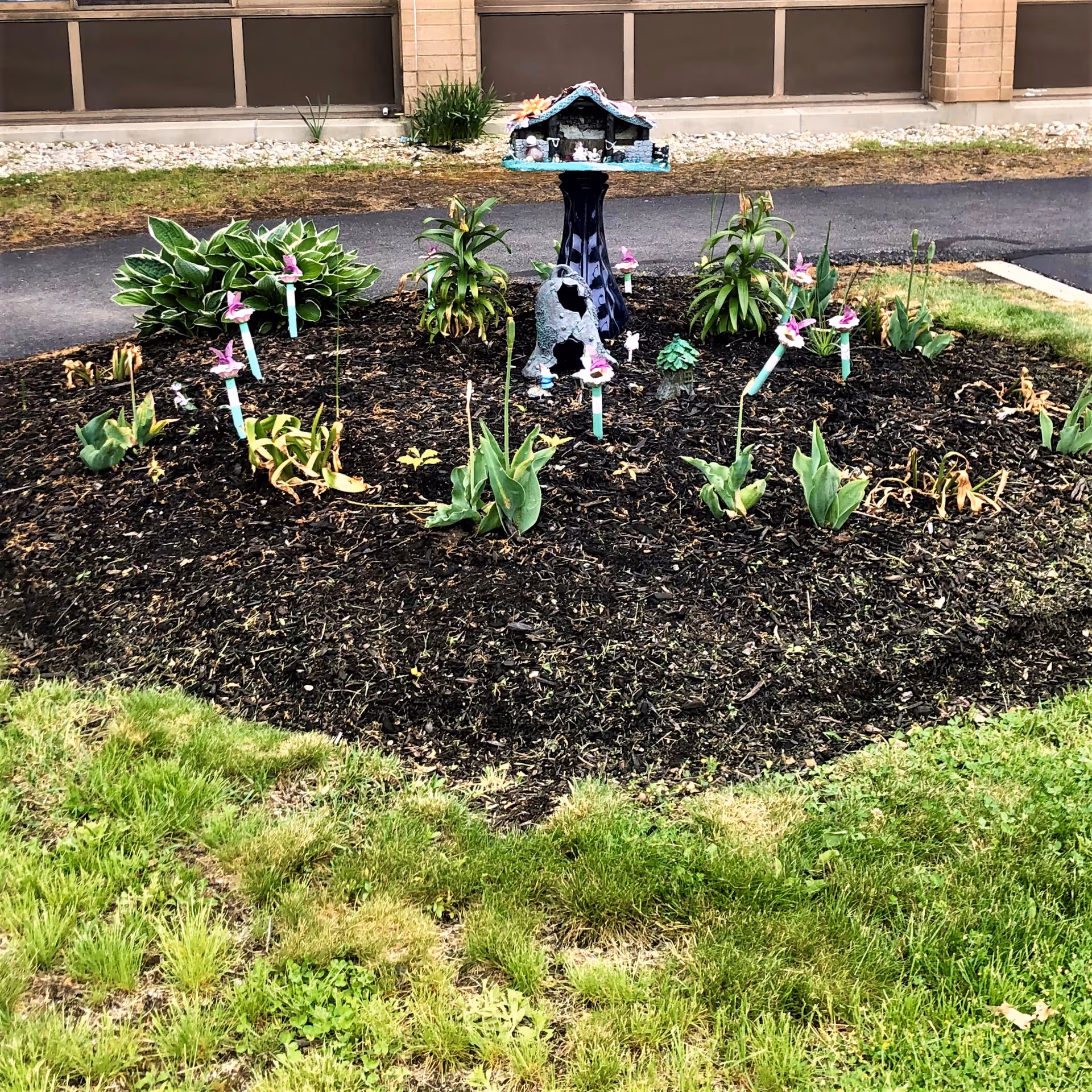 A small circular garden bed with various plants and flowers, some with pink blooms, surrounded by green grass. In the center of the garden bed is a decorative birdhouse mounted on a dark pedestal. The background shows a paved path and the exterior wall of a building with windows.