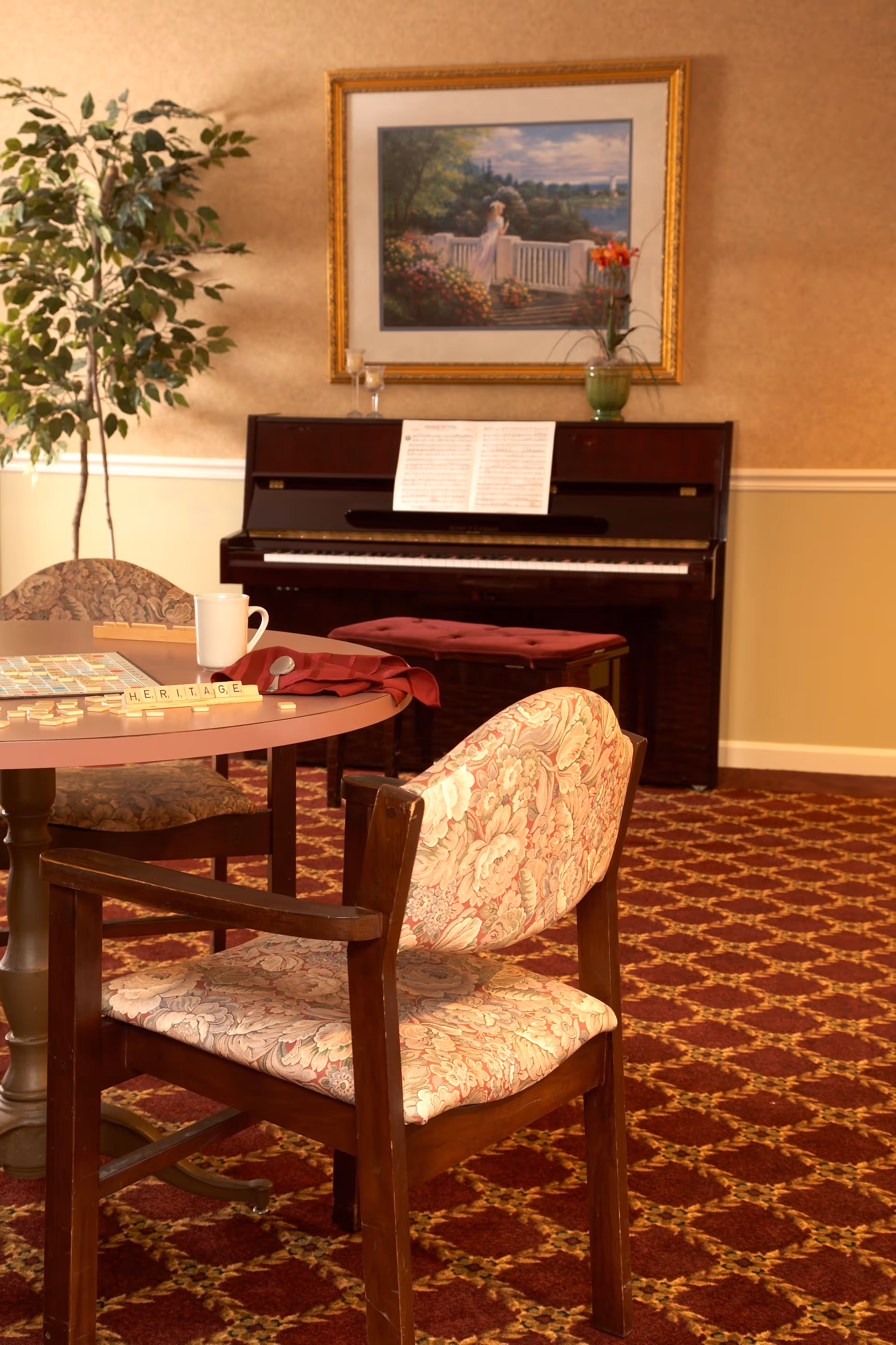 A cozy senior community room with a round table and floral upholstered chairs. On the table, there is a Scrabble game in progress, a white mug, and a folded red cloth. Behind the table, there is an upright piano with sheet music and a cushioned bench. A framed painting of a woman standing on a balcony overlooking a garden and water hangs on the wall above the piano. A potted plant is placed to the left of the piano. The room has patterned carpet and warm beige walls with white trim.