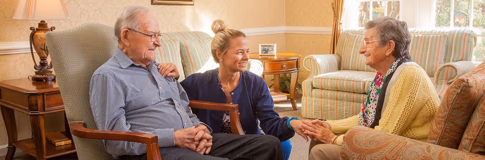 An elderly man and woman sitting in a cozy living room with a caregiver kneeling between them, holding the elderly woman's hands and smiling. The room has comfortable armchairs, a side table with a lamp, and a window with curtains letting in natural light.