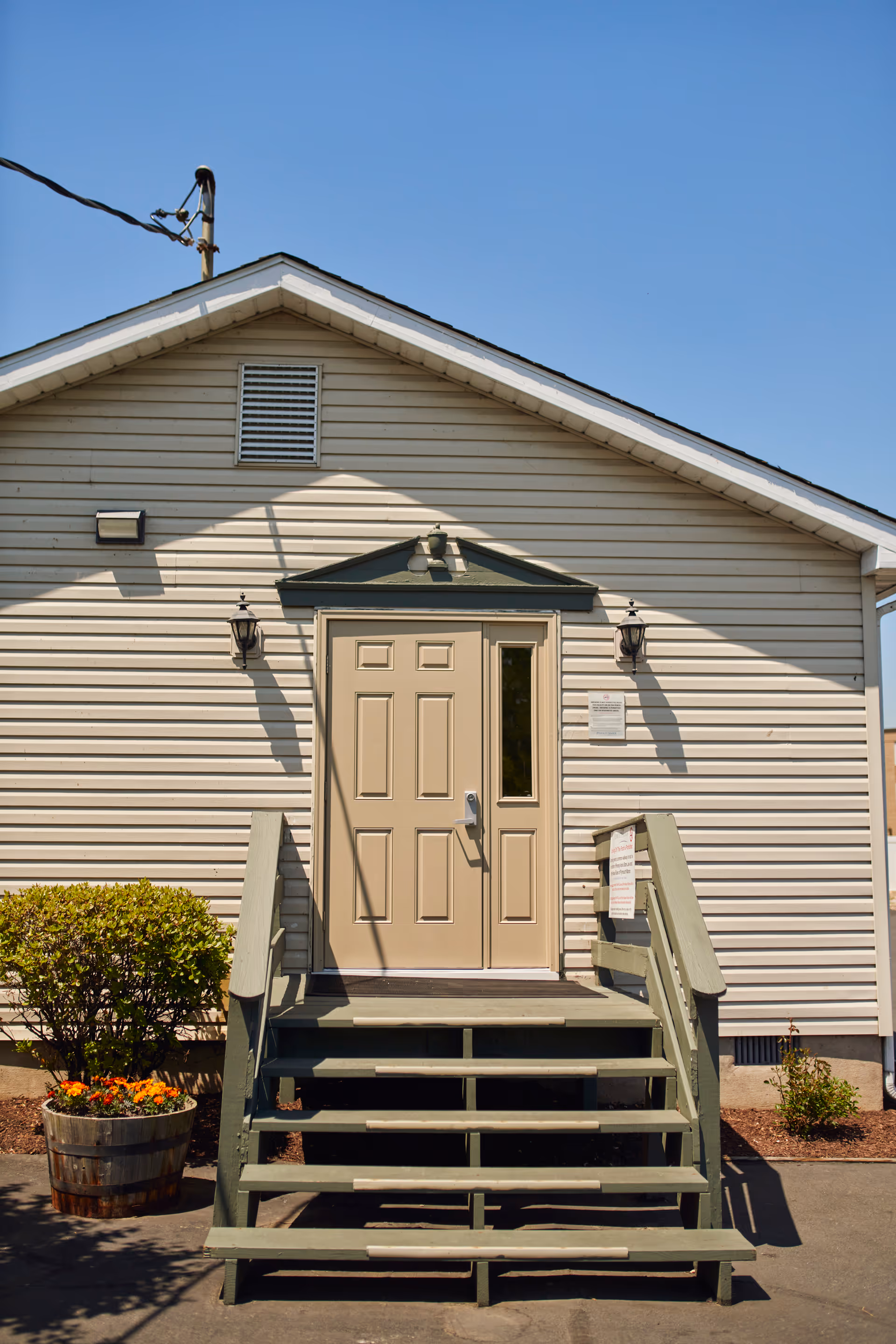 Exterior view of a beige building with a small staircase leading up to a closed door. The building has horizontal siding, two wall-mounted lantern lights on either side of the door, and a small bush with flowers in a wooden planter to the left of the stairs. The sky is clear and blue.