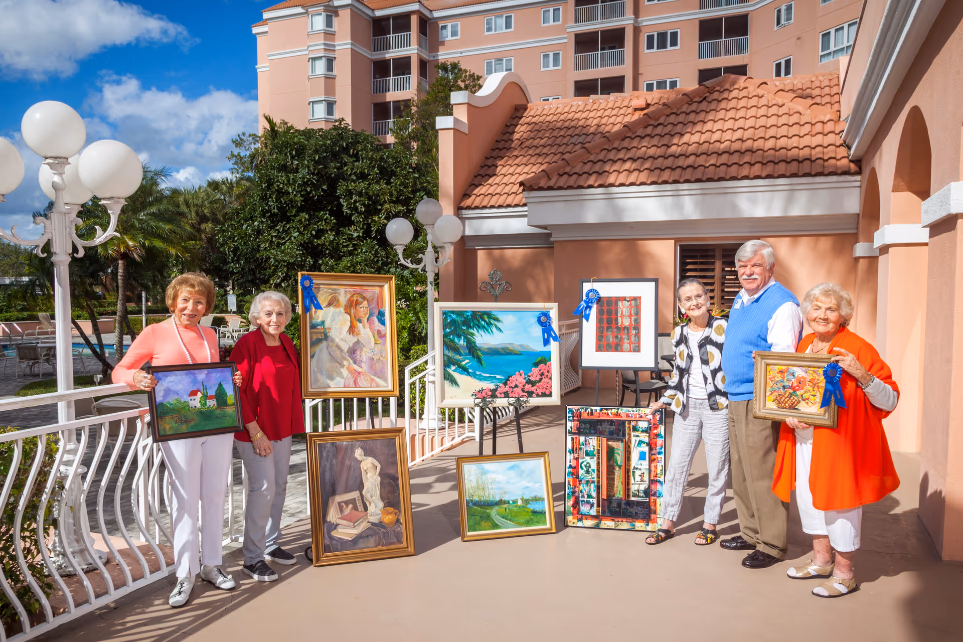 A group of six elderly people standing outside on a sunny patio, each holding or standing near framed paintings, some with blue ribbons indicating awards. The background shows a peach-colored building with a tiled roof and a multi-story residential building behind it.