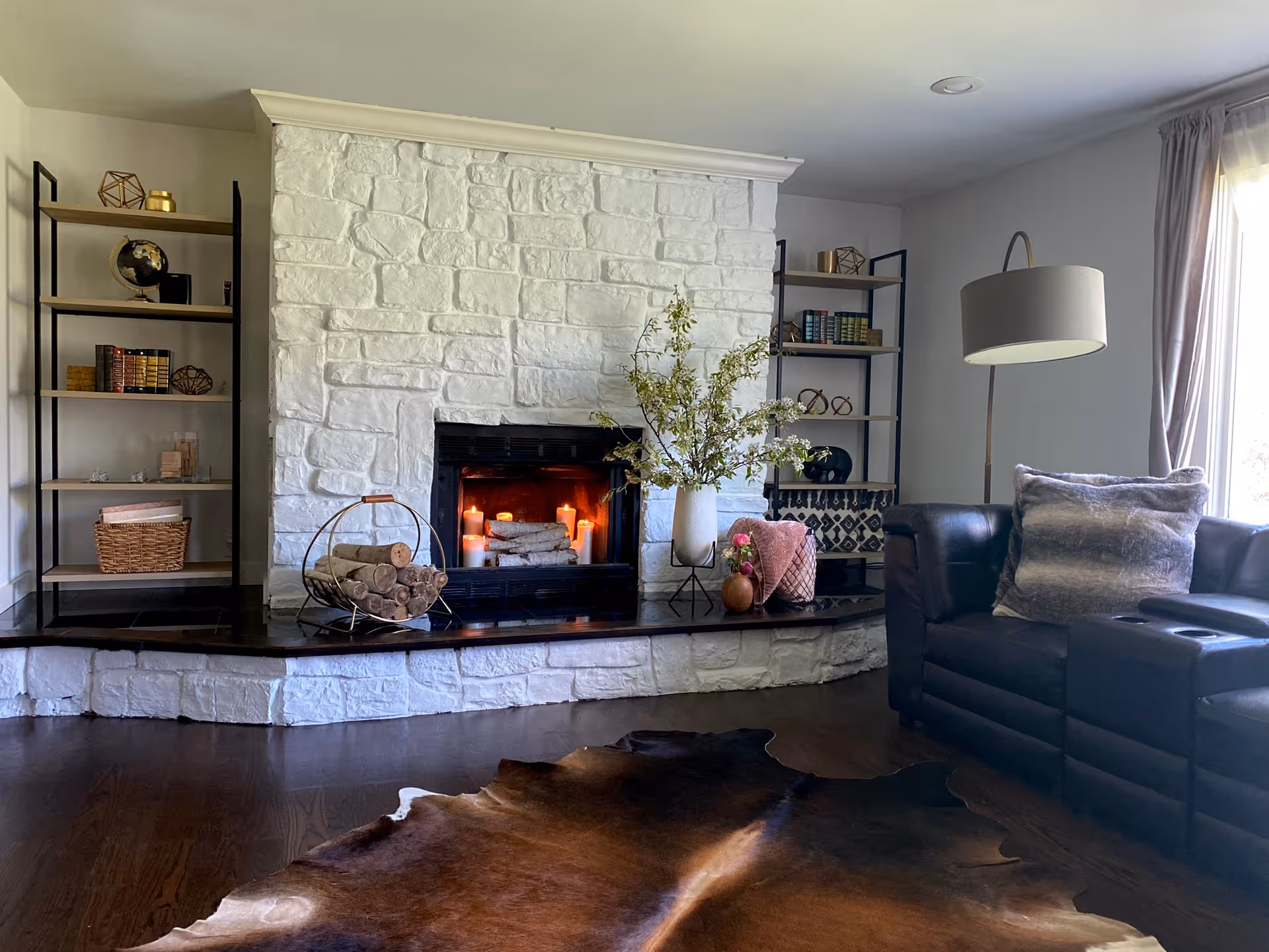 A cozy living room with a white stone fireplace in the center, decorated with candles and logs inside. On either side of the fireplace are black metal and wood shelves holding books, decorative items, and baskets. A black leather sofa with a gray pillow is positioned to the right, next to a large window with light curtains. A floor lamp with a beige shade stands beside the sofa, and a brown animal hide rug lies on the wooden floor in front of the fireplace.