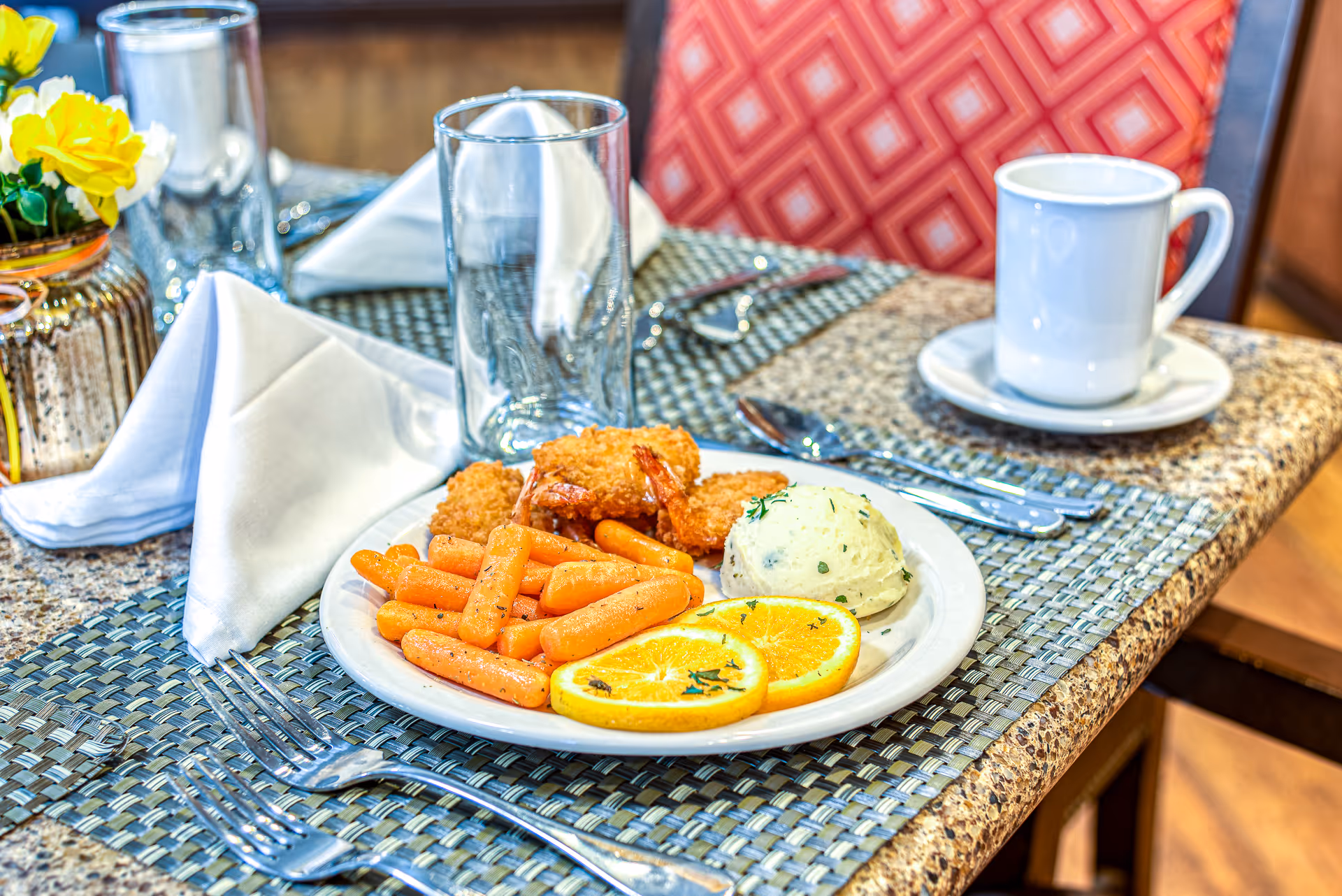 A dining table set with a plate of food including cooked baby carrots, breaded fried shrimp, mashed potatoes garnished with herbs, and two slices of orange. The table has a woven placemat, silverware, a white napkin, a glass, a white coffee cup on a saucer, and a small vase with yellow flowers. The background shows a red patterned chair.
