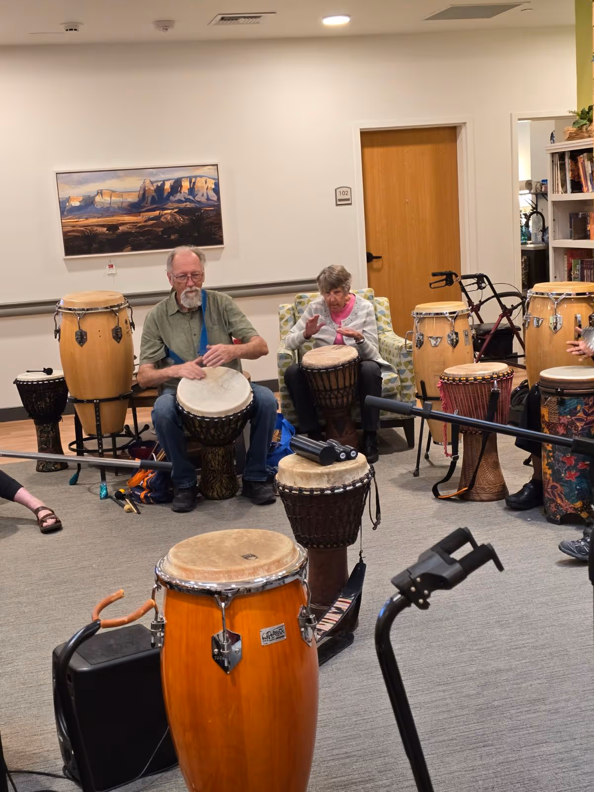 Two elderly individuals seated in a room playing various drums, including djembes and congas. The room has a light-colored wall with a landscape painting, a wooden door labeled 102, and a bookshelf. Several drums are arranged around the participants, and a walker is visible near the door.