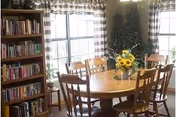 Sunlit dining room with a round wooden table surrounded by chairs, a vase of sunflowers, a bookshelf, and large windows.