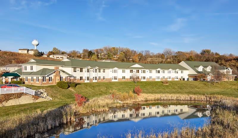 Wide exterior view of Meadow Lakes Senior Living facility with a large two-story building featuring green roofs and white walls. The building is situated on a grassy hill with a pond in the foreground reflecting the structure. Trees and a water tower are visible in the background under a clear blue sky.