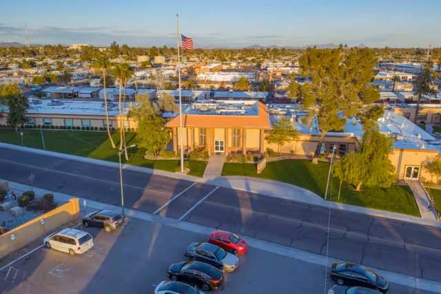 Aerial view of Sunview Health And Rehabilitation Center showing a single-story building with a flat roof, an American flag on a flagpole in front, surrounded by green lawns and trees. Several cars are parked in a parking lot across the street from the building. The surrounding area includes other buildings and palm trees under a clear sky.