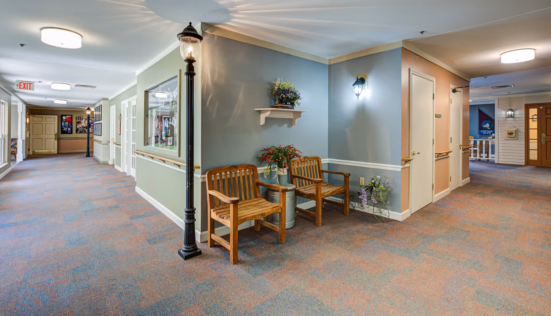 A well-lit indoor hallway in a senior living facility with carpeted floors and pastel-colored walls. Two wooden chairs with a small table and decorative plants are placed in a corner. The hallway features handrails along the walls, framed pictures, and a streetlamp-style light fixture. Doors and exit signs are visible along the corridor.