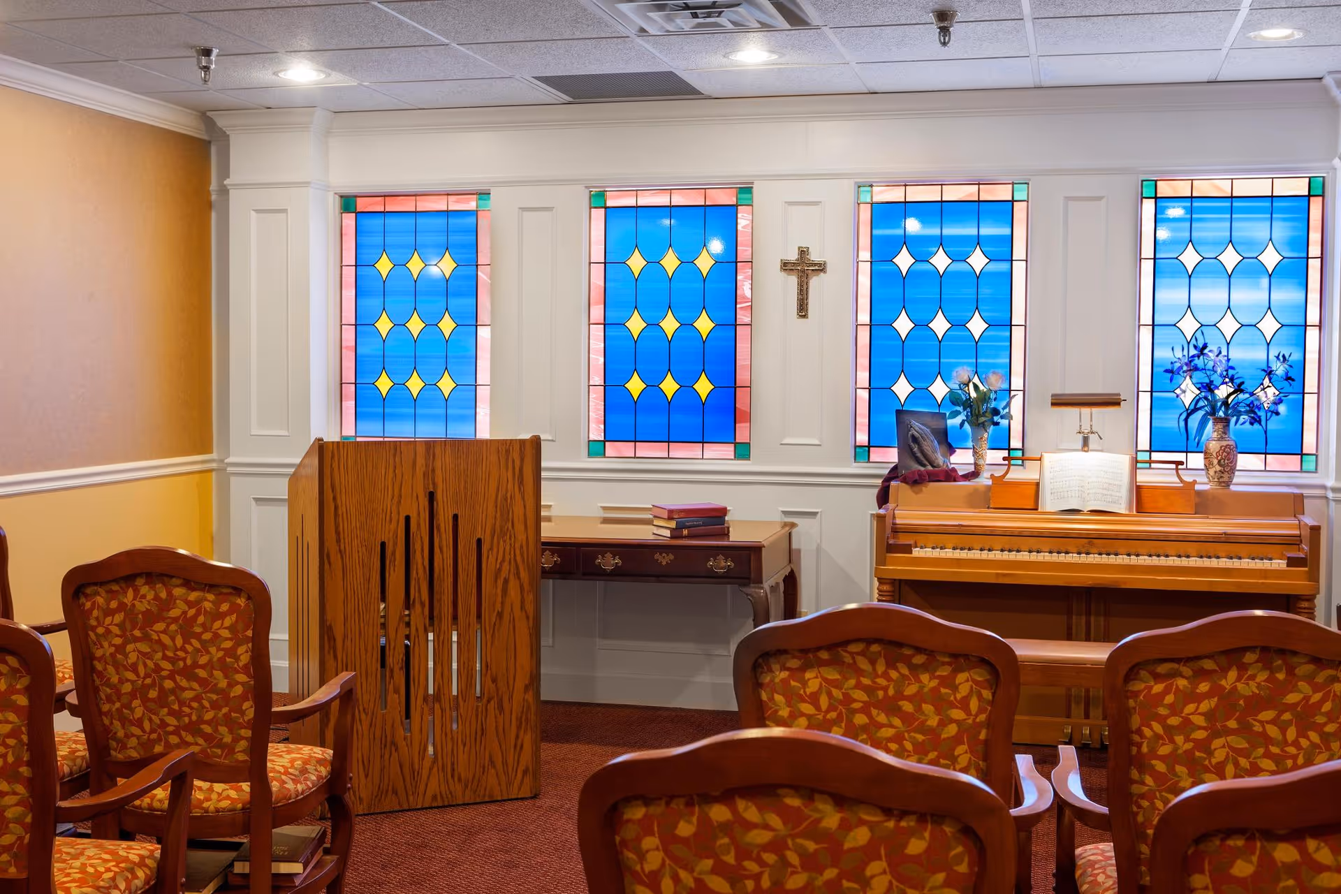 Interior chapel-style room with stained-glass windows, a wooden piano, lectern, and rows of patterned chairs.