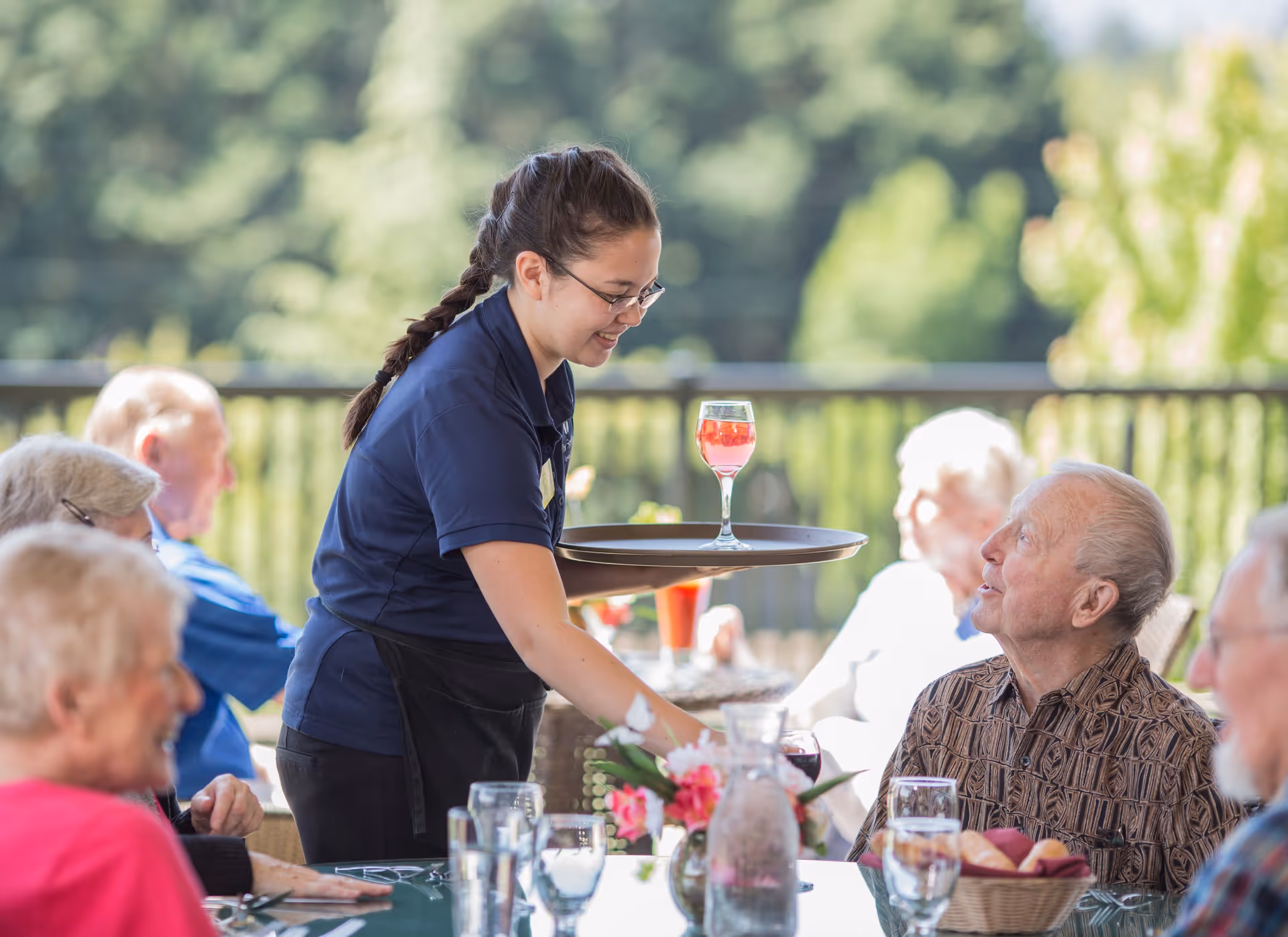 A server hands a drink to elderly residents seated at an outdoor dining table on a sunny patio.