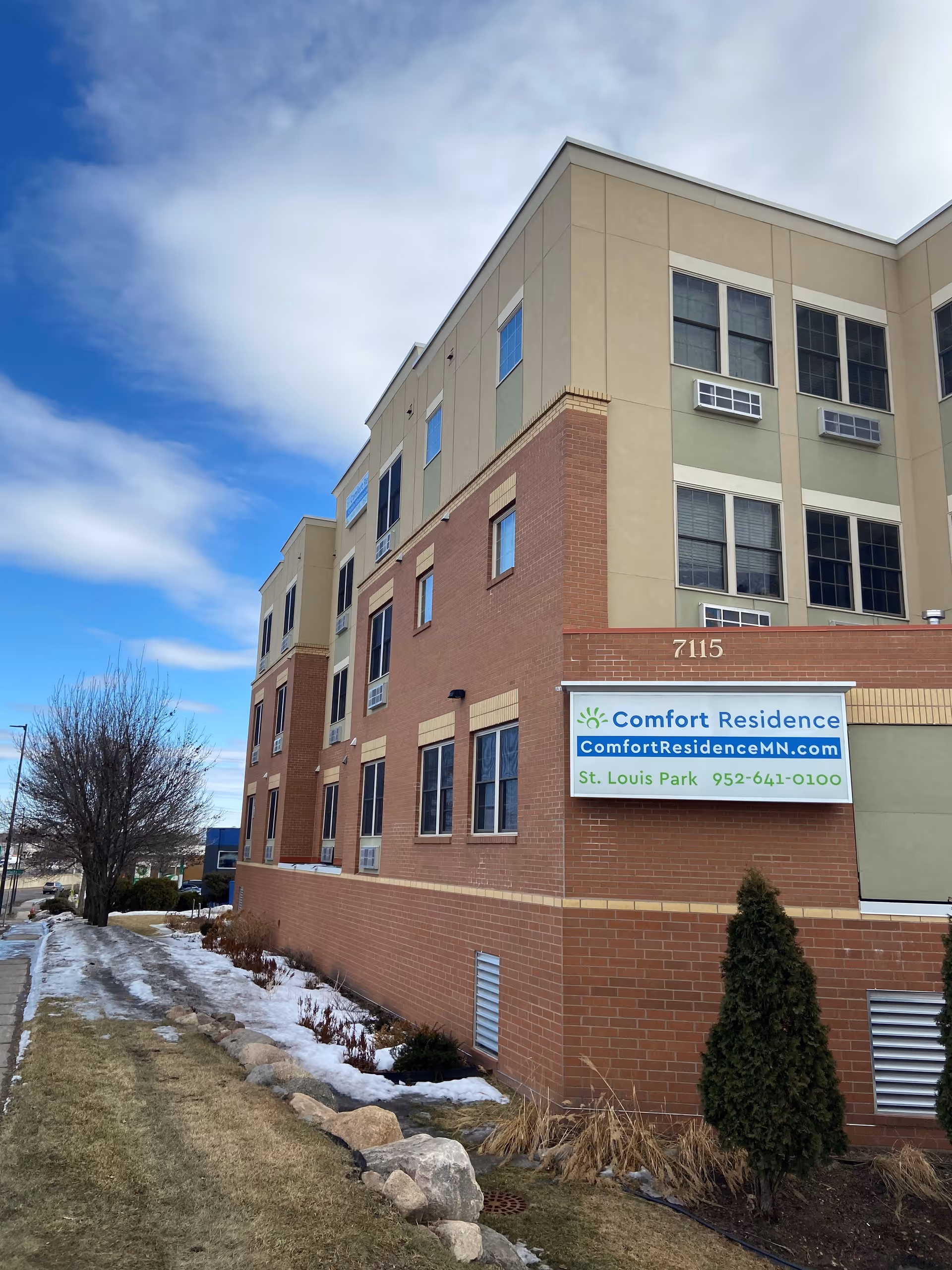 Front exterior of a multi-story senior living building with a 'Comfort Residence' sign and address number 7115.