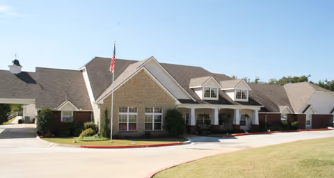 Exterior view of a single-story senior living facility building with a stone and brick facade, multiple windows, a covered entrance, and an American flag on a flagpole in front. The driveway curves around the front of the building with well-maintained landscaping and a clear blue sky above.