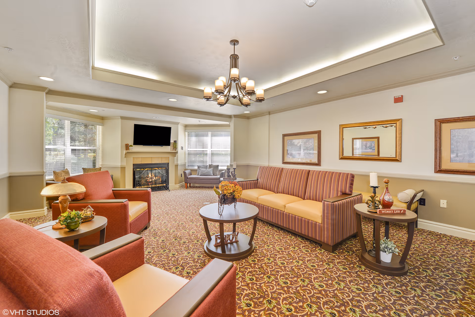 Bright communal living room with a striped sofa, armchairs, coffee tables, a fireplace and wall-mounted TV.