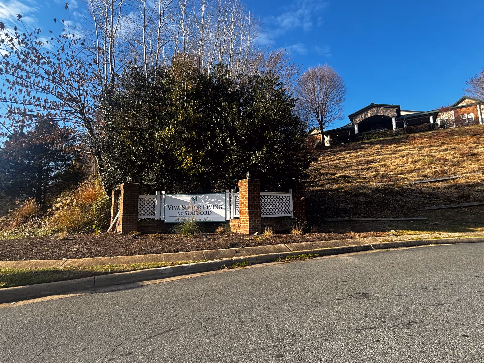 Outdoor view of a senior living facility entrance sign that reads 'Viva Senior Living at Stafford' with a large bush and trees behind it, a clear blue sky, and a building visible on a hill in the background.