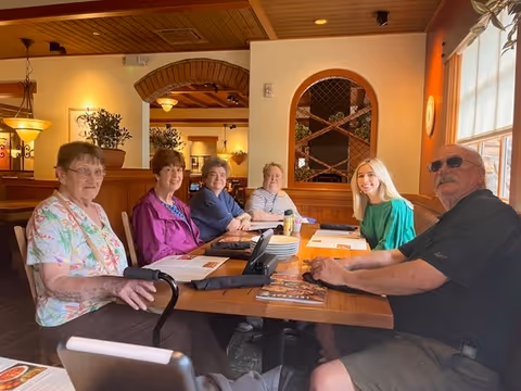 Six people seated around a wooden dining table in a well-lit dining area with menus, plates, and large windows.