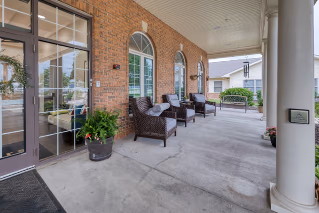 Covered outdoor patio area at Cedarhurst Senior Living of Granite City with wicker chairs and cushions arranged along a brick wall with arched windows. There are potted plants near the entrance door and a bench further down the patio. A white column supports the roof of the patio.