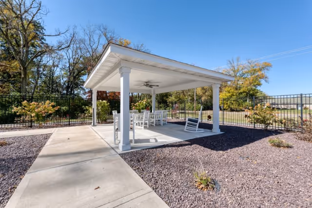 Covered outdoor pavilion with white columns, ceiling fan, table and chairs on a concrete pad surrounded by gravel landscaping and trees.