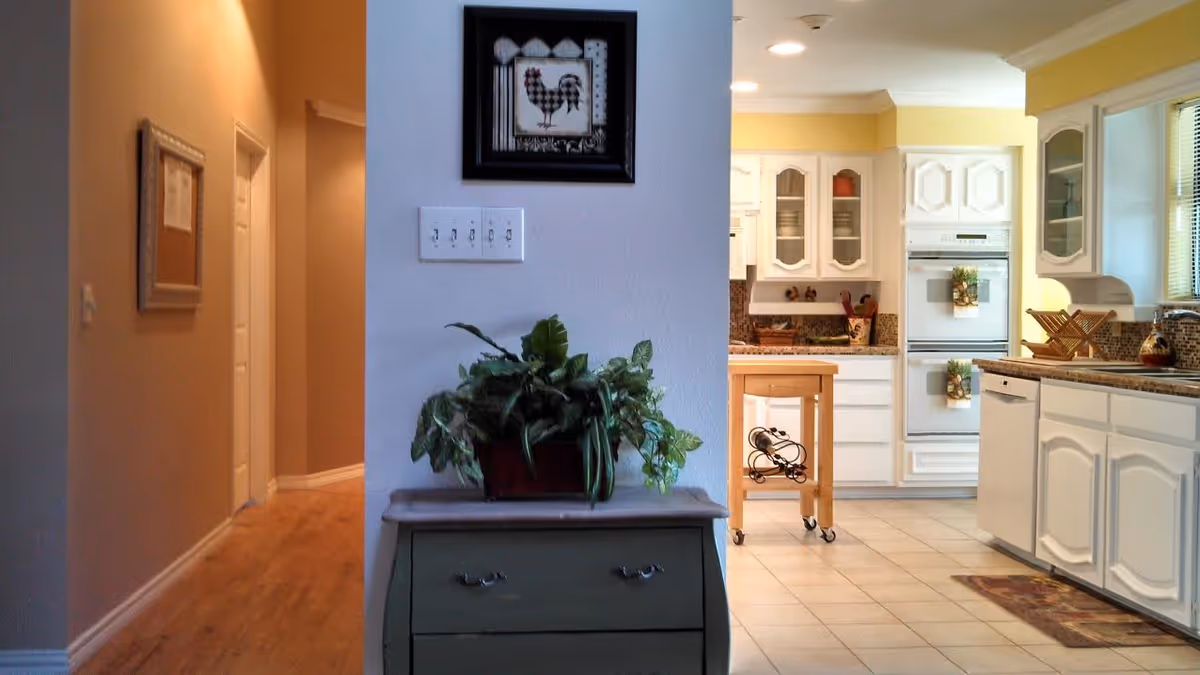 View of a hallway leading to a bright kitchen with white cabinets, a dishwasher, and a small wooden kitchen cart with a wine rack. A green plant sits on a small chest of drawers in the foreground, and a framed picture of a rooster hangs on the wall above the light switches.