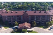 Aerial view of a multi-story brick senior living building with a red roof, central covered entrance and circular driveway, set beside trees and a lake.