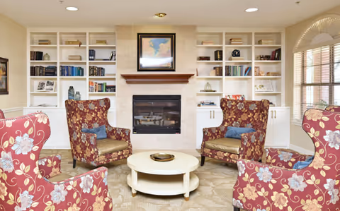 A cozy living room with four floral-patterned armchairs arranged around a round white coffee table. Behind the chairs is a fireplace with a framed picture above it, flanked by built-in white bookshelves filled with books and decorative items. A large window with wooden blinds allows natural light into the room.