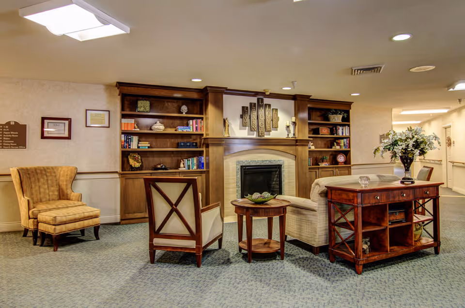 A cozy living room area in Breeze Park facility featuring a fireplace with wooden built-in bookshelves on either side filled with books and decorative items. There are two armchairs, a small round wooden table, and a wooden console table with a vase of flowers. The room has soft lighting and carpeted flooring.
