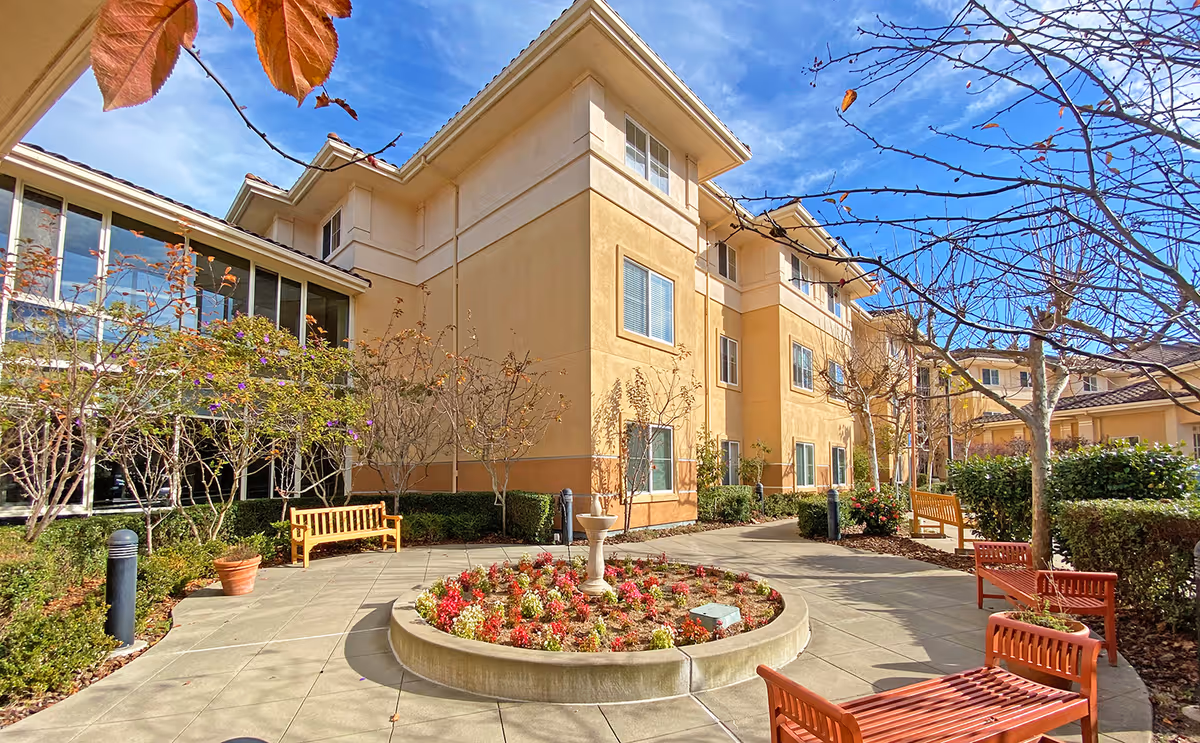 Outdoor courtyard area of a senior living facility with a circular flower bed in the center, surrounded by benches and trees with some autumn leaves. The building is a multi-story structure with beige and light brown walls and several windows. The sky is clear and blue.