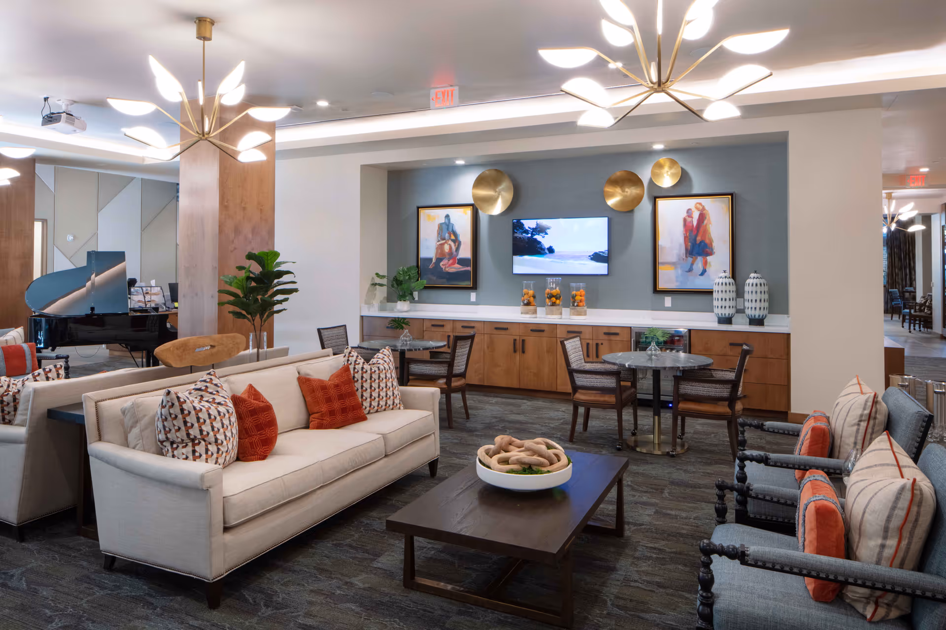 A modern and cozy living room area in a senior living facility featuring a beige sofa with patterned and orange pillows, two blue armchairs with striped and orange pillows, a dark wooden coffee table with a decorative bowl, two small round tables with chairs, a wall-mounted TV, decorative paintings, and brass wall accents. The room is well-lit with contemporary chandeliers and has a grand piano in the background.