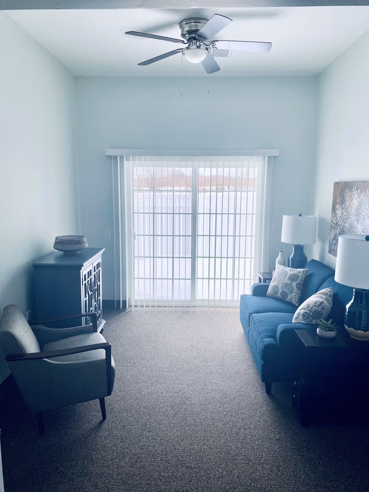 Bright living room with a blue sofa and armchair facing sliding glass doors with vertical blinds, a ceiling fan overhead, and side tables with lamps.
