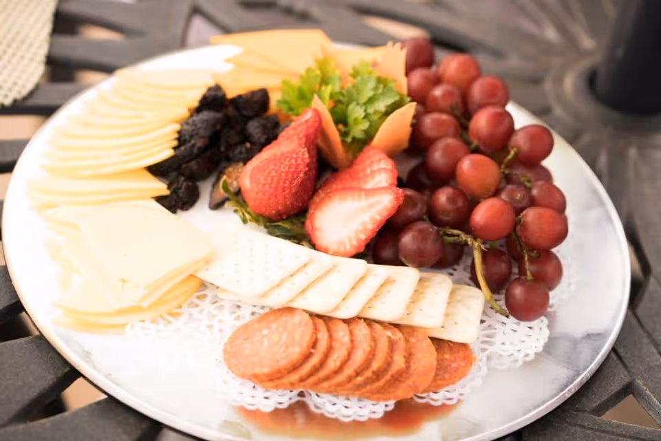 A silver platter with an assortment of snacks including sliced cheese, dried fruit, strawberries, red grapes, crackers, and slices of pepperoni, garnished with parsley and carrot curls, placed on a black metal table.