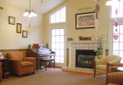 A cozy living room area with a fireplace, a piano, and comfortable armchairs. The fireplace is tiled and decorated with small items and a framed picture above it. There are large windows and a glass door letting in natural light. The room has warm lighting from a ceiling fixture and hanging decorations.