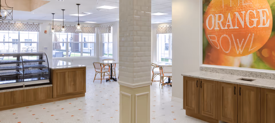 A bright dining area with large windows, small tables with chairs, a display case on the left, and wooden cabinets with a granite countertop on the right. A large colorful wall art featuring an orange with the text 'ORANGE BOWL' is visible above the cabinets.