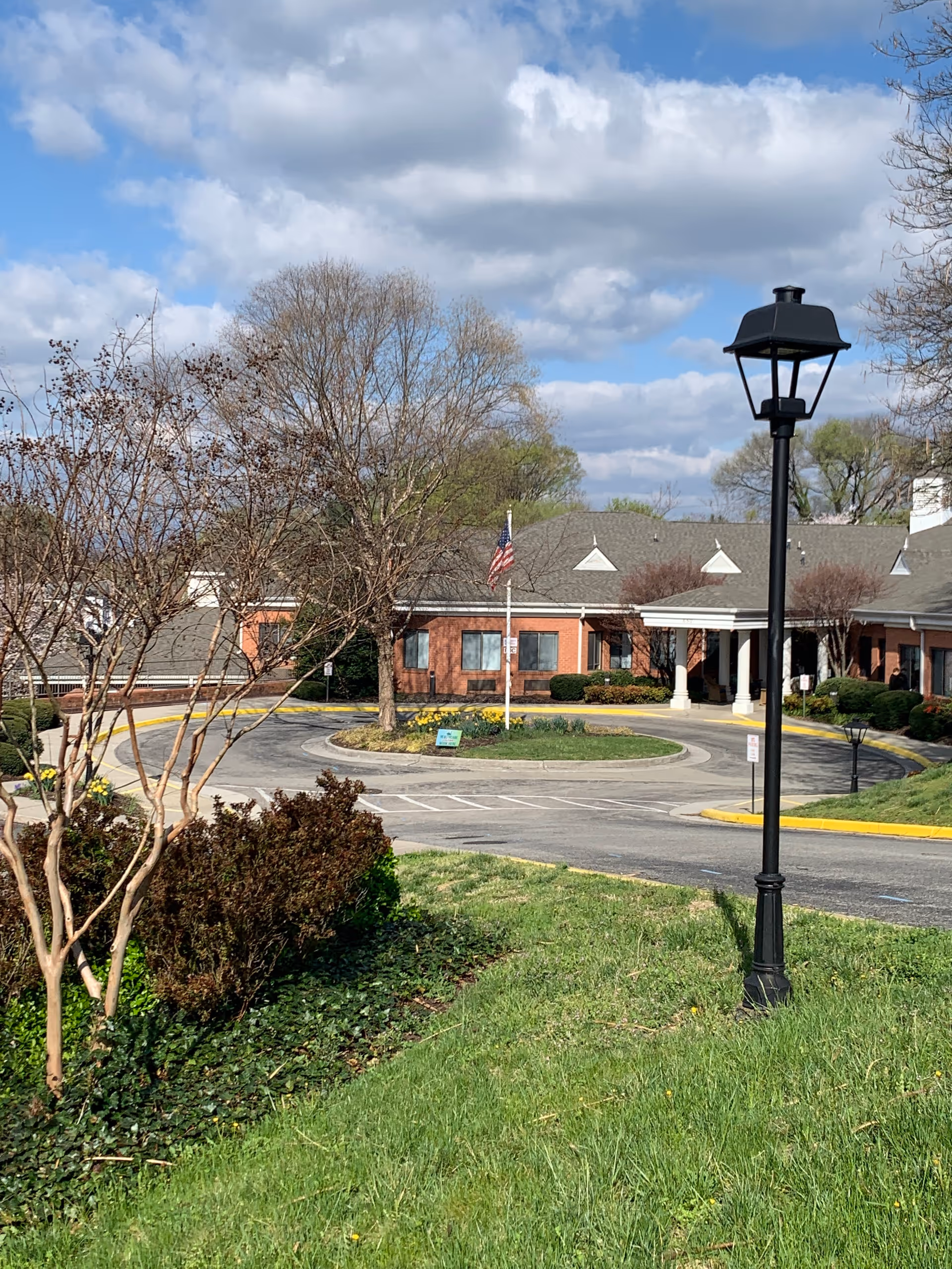 Front exterior of a brick senior living building with a circular driveway, flagpole, lamp post, and landscaped lawn under a partly cloudy sky.