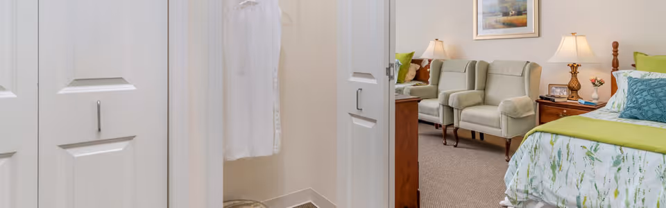 A resident bedroom showing a partially visible bed with green-and-blue bedding, two upholstered chairs and lamps, and an open closet with a hanging garment.