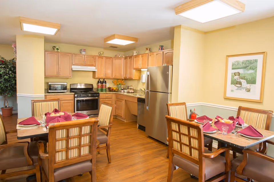 A cozy dining area and kitchen in a senior living facility. The kitchen features wooden cabinets, a stainless steel refrigerator, stove, microwave, and dishwasher. Two dining tables are set with plates, glasses, and burgundy napkins, surrounded by wooden chairs with striped cushions. The walls are painted light yellow with a framed picture and a fire extinguisher visible.