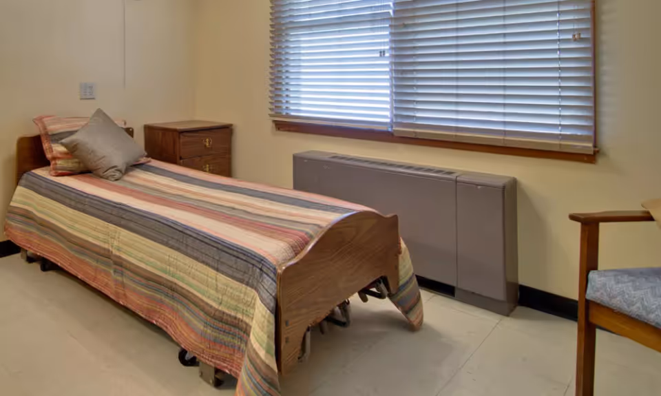 Small bedroom with a single bed covered by a striped bedspread, a nightstand, window blinds, and a chair.