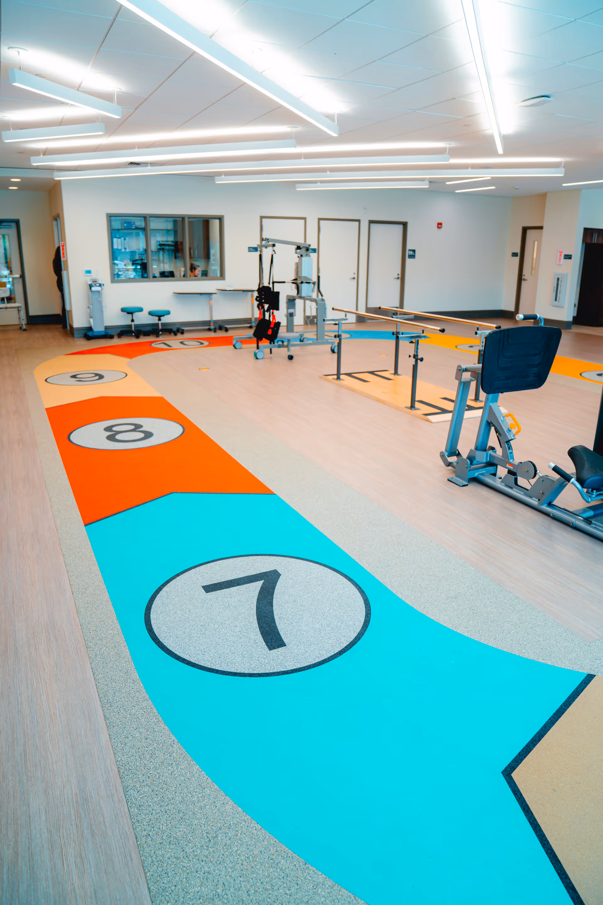 A bright indoor rehabilitation or physical therapy room with colorful numbered floor markings from 7 to 10, parallel bars for walking exercises, and exercise equipment including a seated leg press machine. The room has a light wood floor, white walls, multiple doors, and modern ceiling lights.